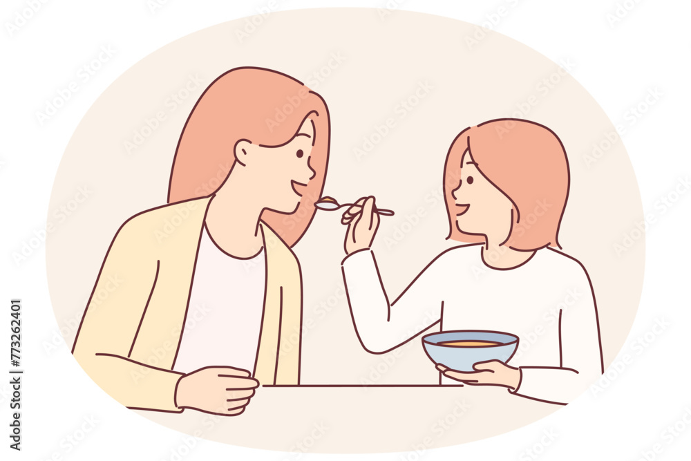 Little girl feeds mother with spoon during breakfast sitting at kitchen table spending time together Little girl feeds mother with spoon during breakfast sitting at kitchen table spending time together