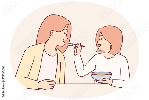 Little girl feeds mother with spoon during breakfast sitting at kitchen table spending time together