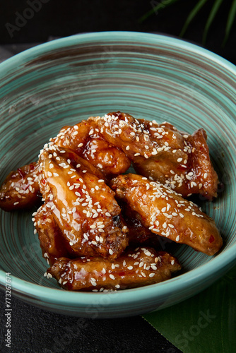 Roasted chicken wings in teriyaki sauce with sesame on ceramic bowl. Buffalo wings in asian style on black concrete background. Aesthetic composition with fried chicken wings, chopsticks and leaf.