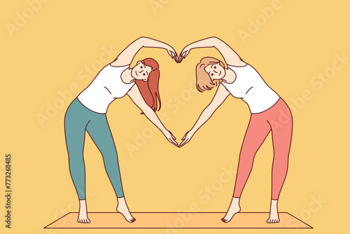 Paired yoga of two women doing aerobics or pilates standing on sports mat and making heart sign from hands. Attractive girls are interested in fitness and pilates to avoid excess weight