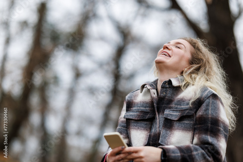 Woman Laughing While Holding Cell Phone in a Forest During Fall