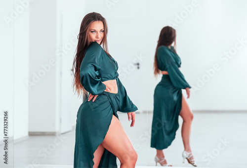 Graceful woman dressed in dark green Latin dancing dress doing elegant dance pas in white color big hall with big mirror wall. People's expressions during dancing, beauty of woman's body concept image