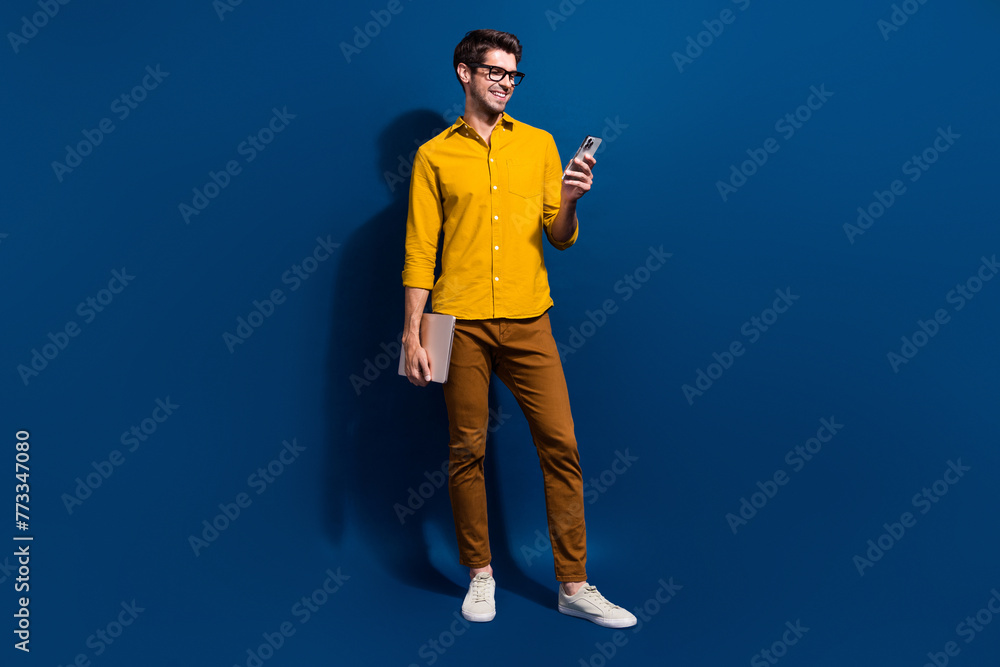 Full size photo of handsome young man hold netbook device smm manager dressed stylish yellow outfit isolated on dark blue color background Full size photo of handsome young man hold netbook device smm manager dressed stylish yellow outfit isolated on dark blue color background