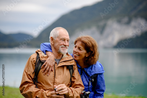 Portrait of beautiful active elderly couple hiking together in autumn mountains. Senior tourists embracing each other in front of lake.