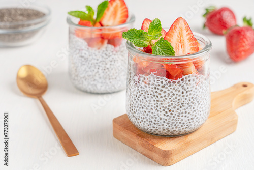 Healthy chia pudding sweet dessert made with seeds soaked in vegetarian milk with chopped strawberry topping and fresh mint leaf served in glass jar on chopping board on white wooden table with spoon