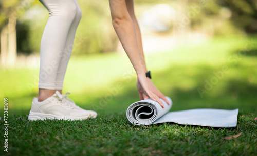 Close-up of a person in white leggings and sneakers rolling up a yoga mat in a lush green park