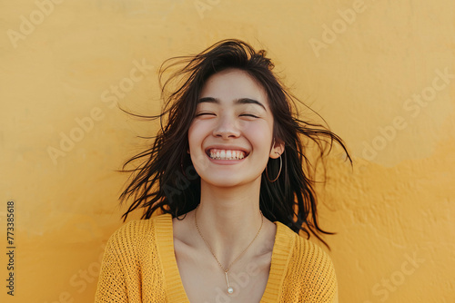 Joyful young woman smiling with hair blowing in the wind, against a yellow textured wall.