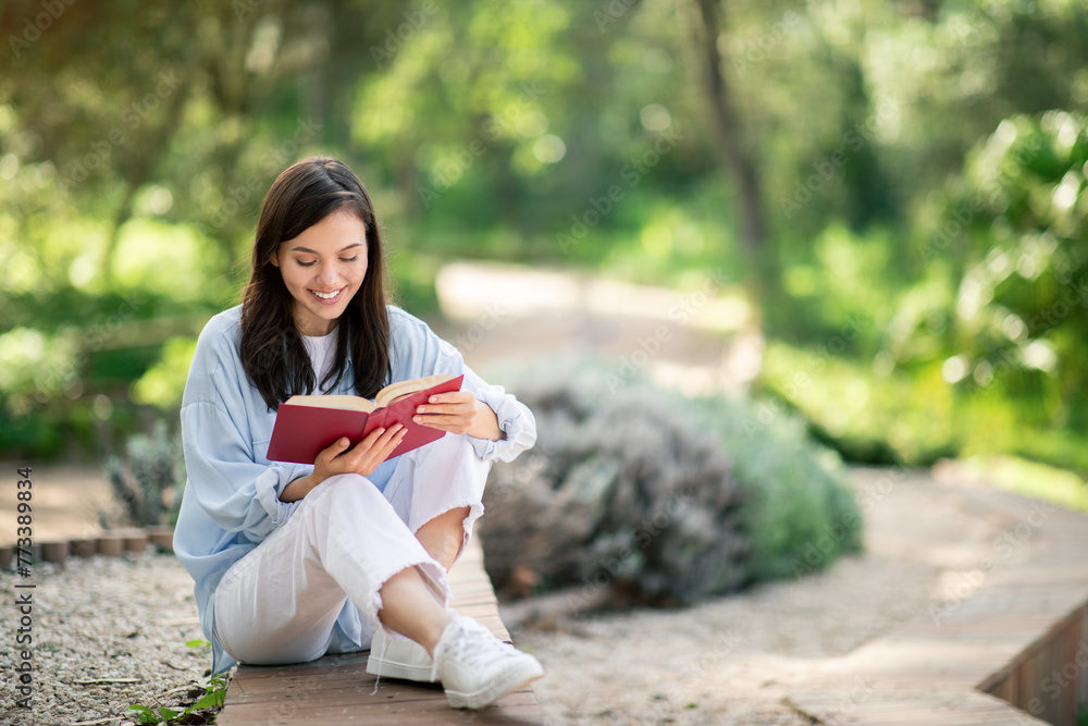 Contented young woman deeply engrossed in reading a red book, sitting with legs crossed on a park path Contented young woman deeply engrossed in reading a red book, sitting with legs crossed on a park path