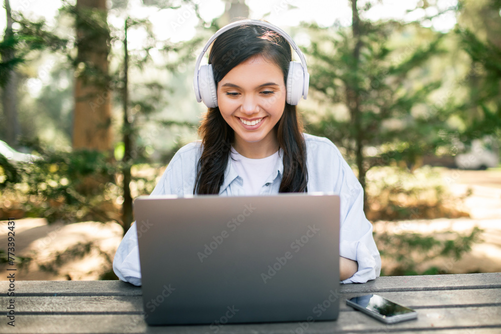 A young woman with headphones smiles while using a laptop at an outdoor wooden table A young woman with headphones smiles while using a laptop at an outdoor wooden table