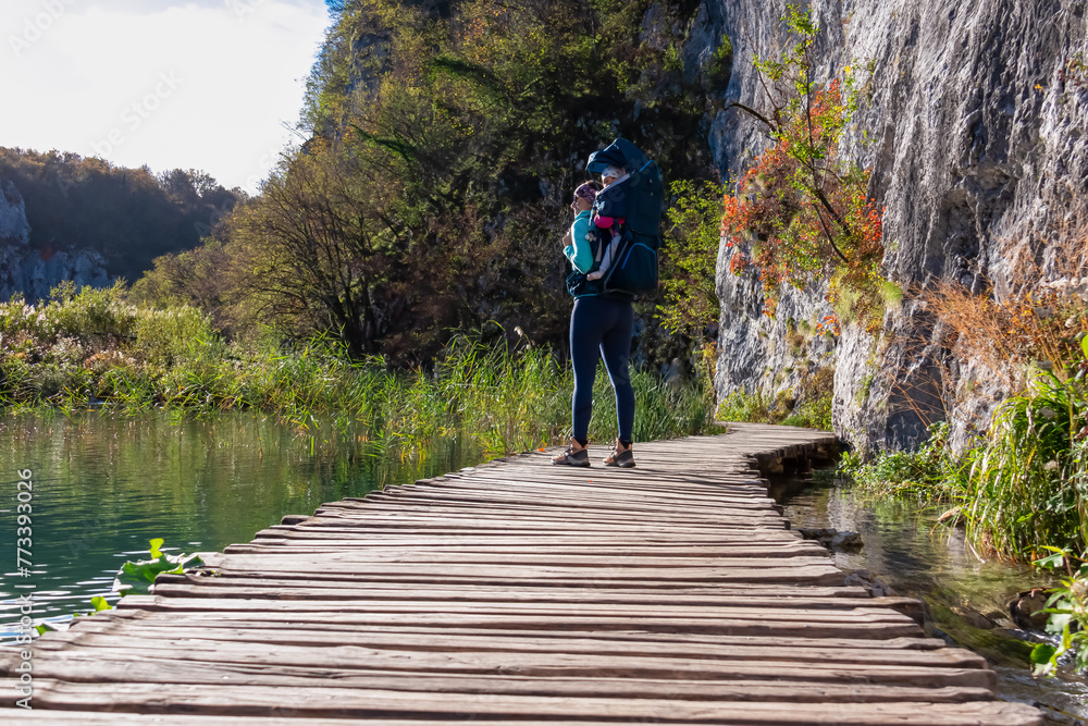 Hiker woman with baby carrier on enchanting hiking trail on rustical wooden footpath in Plitvice lakes National Park, Karlovac, Croatia, Europe. Walking over turquoise water in idyllic paradise nature