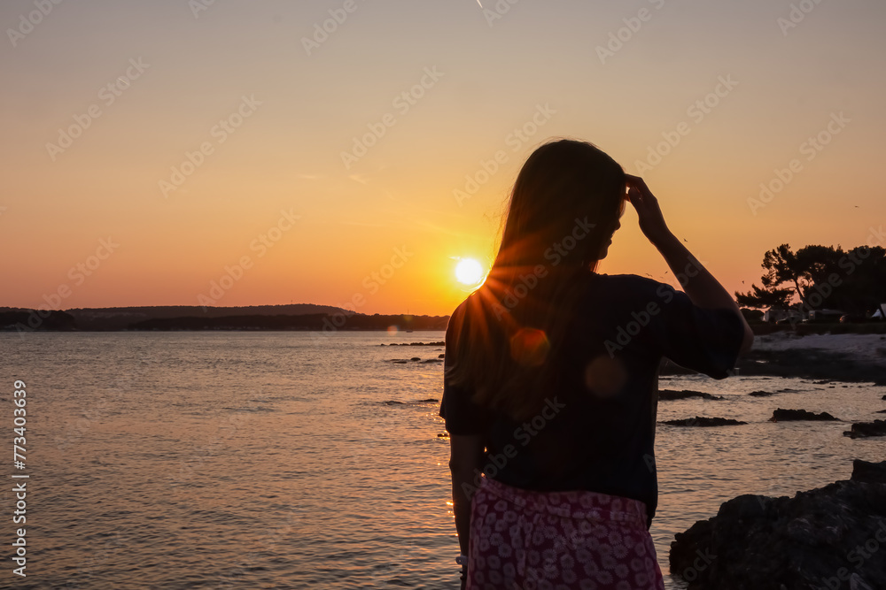 Fototapeta premium Silhouette of woman with on idyllic beach at romantic sunset in Medulin, Istria peninsula, Croatia. Sunglitter on calm water surface of Adriatic Mediterranean Sea, Kvarner Gulf in summer. Dramatic sky