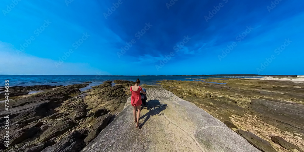 Fototapeta premium Loving mother walking with baby stroller along coastal road in Medulin, Istria peninsula, Croatia, Europe. Idyllic coastline of Kvarner Gulf in Adriatic Mediterranean Sea. Travel destination in summer