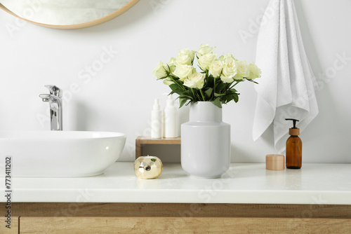 Vase with beautiful white roses and toiletries near sink in bathroom