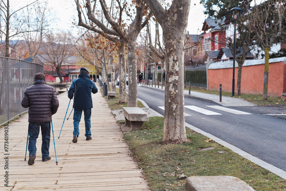 Two men are walking down a sidewalk with the walking sticks or trekking poles. The sidewalk is lined with trees and there is a bench on the sidewalk. The street is empty and there are no other people Two men are walking down a sidewalk with the walking sticks or trekking poles. The sidewalk is lined with trees and there is a bench on the sidewalk. The street is empty and there are no other people