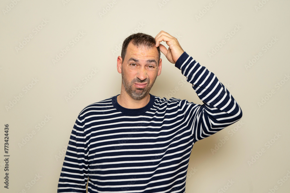 Bearded Hispanic man in his 40s wearing a striped sweater scratching his head while trying to make a decision, doubt concept. Isolated on beige studio background. Bearded Hispanic man in his 40s wearing a striped sweater scratching his head while trying to make a decision, doubt concept. Isolated on beige studio background.