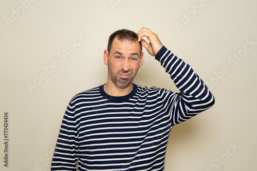 Bearded Hispanic man in his 40s wearing a striped sweater scratching his head while trying to make a decision, doubt concept. Isolated on beige studio background.