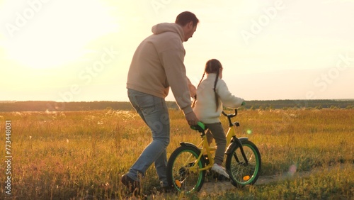 Parent teaches child to ride bicycle. Father and daughter in field at epic sunset. Child safety, dad regulates and controls beloved girl kid on bike, warning against injuries and bruises. Slow motion