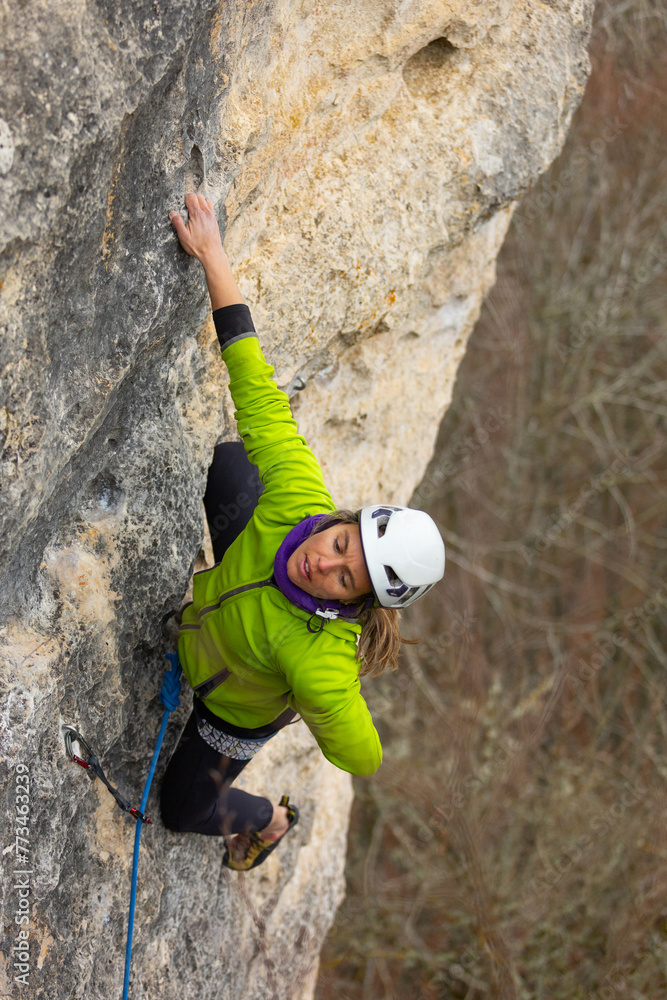 A woman is climbing a rock wall in a green jacket. She is wearing a helmet and a purple scarf A woman is climbing a rock wall in a green jacket. She is wearing a helmet and a purple scarf