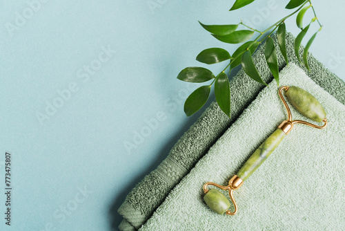 Green jade face roller and green leaves on light blue background. Massage tool for facial skin care, SPA self and beauty care treatment concept.