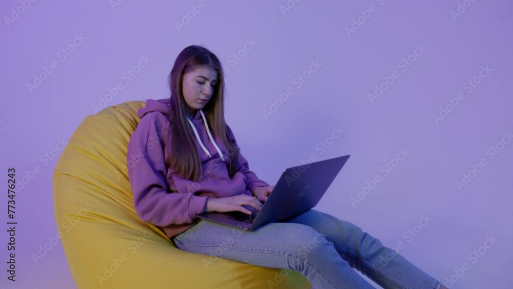 Young woman sitting on pouf and using notebook isolated on purple color background. Girl in hoodie working remote and lying on bean bag. Female programmer trying to fix bug in code and sitting in Young woman sitting on pouf and using notebook isolated on purple color background. Girl in hoodie working remote and lying on bean bag. Female programmer trying to fix bug in code and sitting in