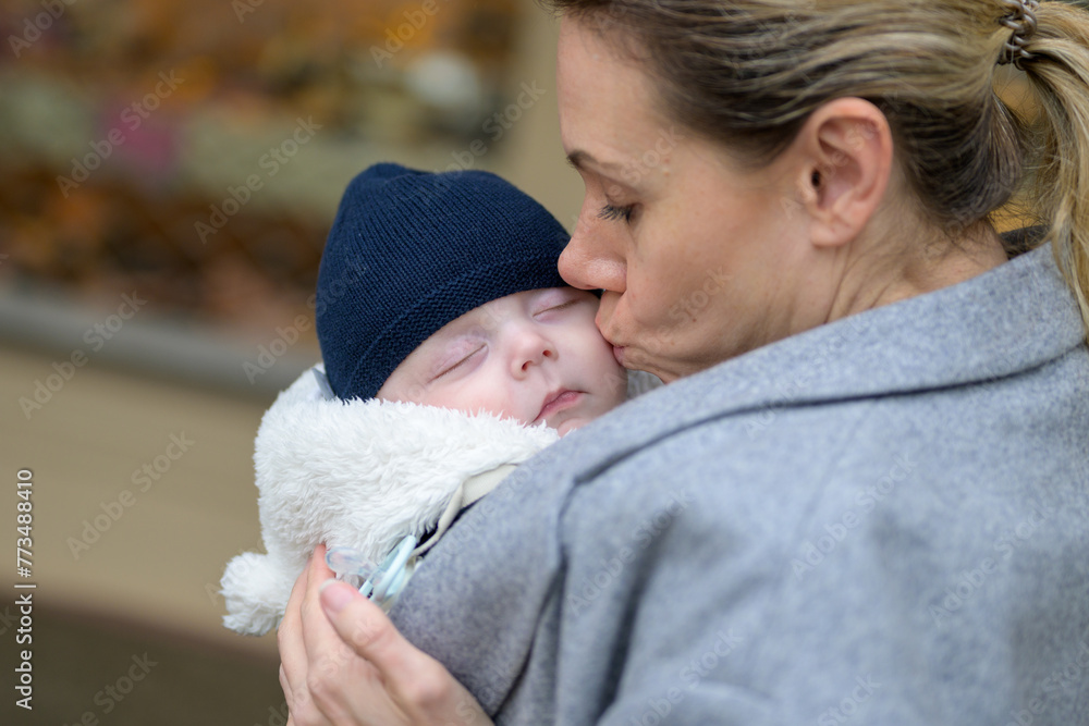 Close up of a happy mother in her 40s holding her newborn baby in a baby carrier and kissing it lovingly Close up of a happy mother in her 40s holding her newborn baby in a baby carrier and kissing it lovingly
