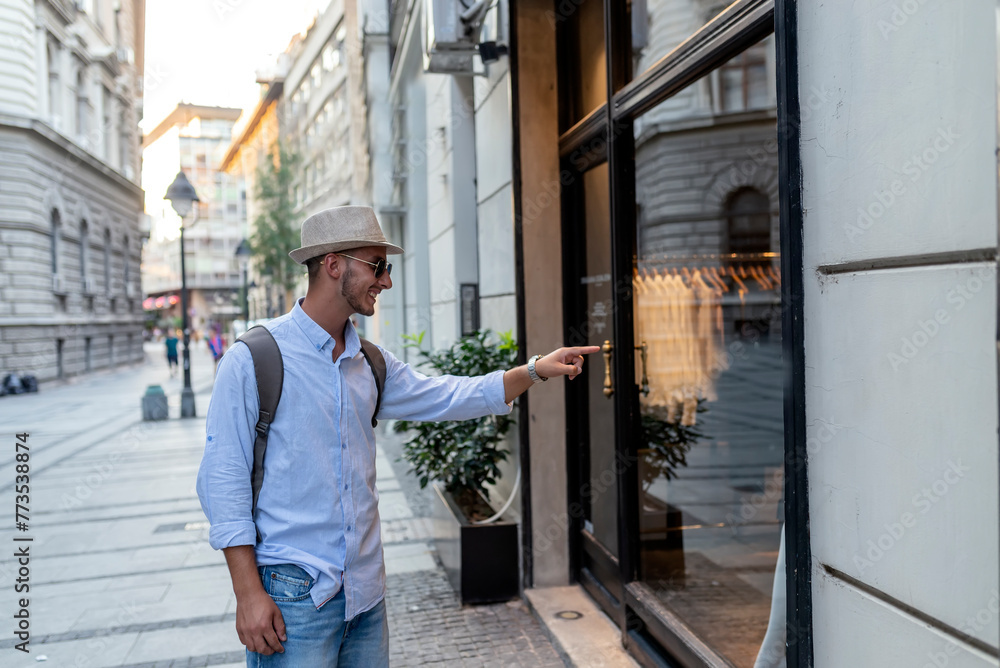 Handsome Well Dressed Young Man with Hat, Sunglasses and Backpack Standing in the City Center and Pointing at the Shopping Window Handsome Well Dressed Young Man with Hat, Sunglasses and Backpack Standing in the City Center and Pointing at the Shopping Window