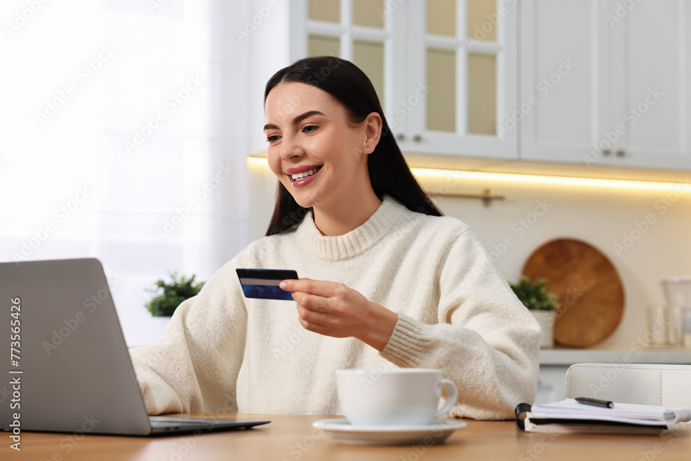 Happy young woman with credit card using laptop for shopping online at wooden table in kitchen Happy young woman with credit card using laptop for shopping online at wooden table in kitchen