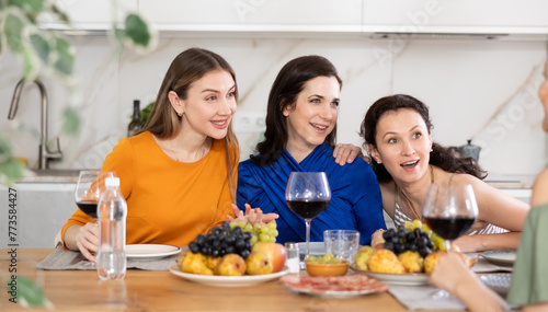 Relaxed adult female friends having fun at intimate house party  sharing stories and laughing gathered around table with wine and appetizers in cozy kitchen