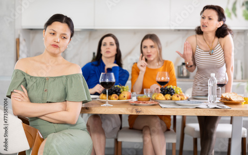 At home party, young Asian woman appearing offended and upset during argument while female friends in background showing disapproval and discontent..