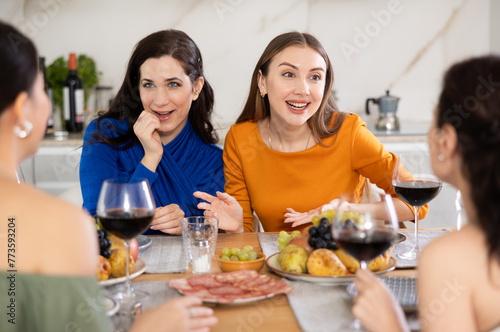 Relaxed adult female friends having fun at intimate house party  sharing stories and laughing gathered around table with wine and appetizers in cozy kitchen