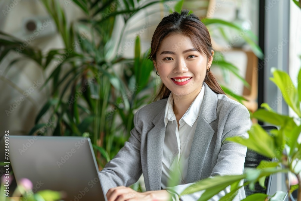 beautiful Asian woman in professional attire is sitting at her desk, smiling and typing on the laptop computer with green plants all around and behind it. beautiful Asian woman in professional attire is sitting at her desk, smiling and typing on the laptop computer with green plants all around and behind it.