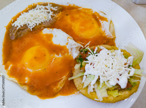 A plate of eggs rancheros with spicy tomato sauce, a sope with vegetables and beans with shredded cheese on top. Mexican breakfast