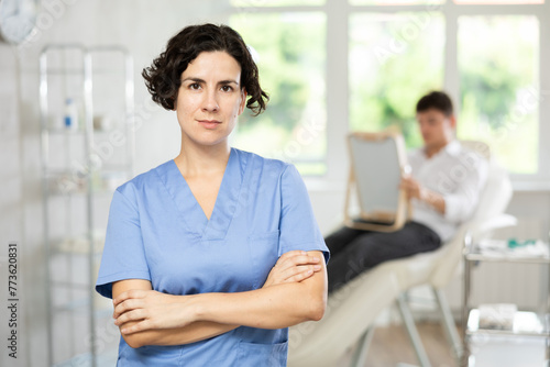 Positive middle-aged woman doctor posing against background of doctor's cabinet with patient waiting for consultation