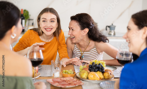 Relaxed adult female friends having fun at intimate house party, sharing stories and laughing gathered around table with wine and appetizers in cozy kitchen