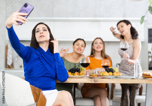 Spanish smiling girl is sitting on chair and taking selfies during gatherings with friends, behind her group of friends are having fun chatting, eating, drinking wine. photo