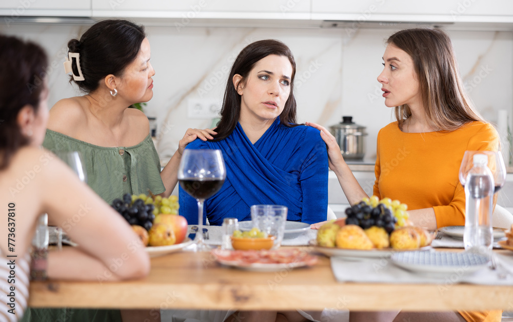 Group of empathetic and caring female friends gathering around table to console upset bestie during friendly get-together with wine at home..