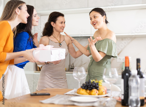 Excited young Asian woman in olive ruffled dress warmly hosting female friends bringing unexpected beautifully wrapped gift for festive home get-together..