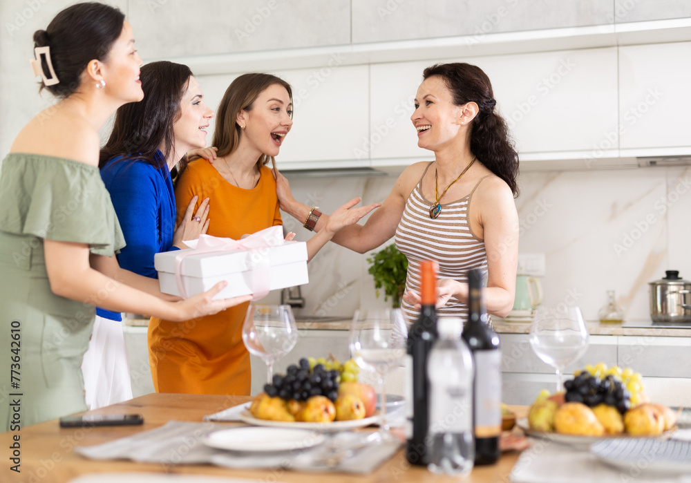 Girls hug and kiss on cheek when they meet, company group of three guests gives gift to birthday woman. Treats, food, alcohol and snacks on table in background. Concept of home party - obrazy, fototapety, plakaty 