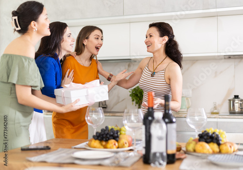 Girls hug and kiss on cheek when they meet, company group of three guests gives gift to birthday woman. Treats, food, alcohol and snacks on table in background. Concept of home party