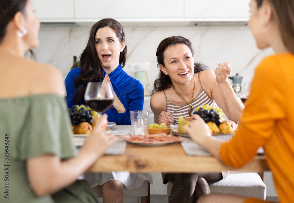 Cheerful female friends enjoying home gathering with wine and light snacks at kitchen table, engaging in lively conversations..