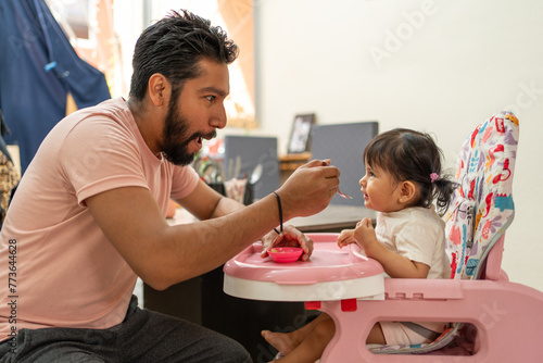 Happy Hispanic father feeding his cheerful baby girl, toddler enjoying meal in high chair, dad and daughter sharing a joyful momen photo