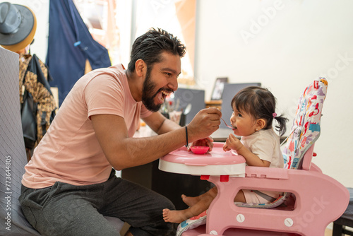 hispanic father feeding her cheerful baby girl, toddler eating in high chair, dad and daughter smiling in a lovely moment photo