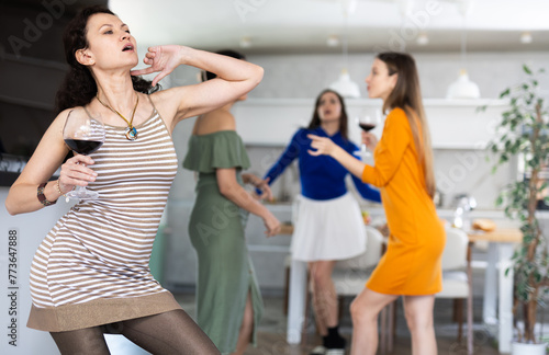 Emotional slightly tipsy woman in striped dress dancing with glass of wine in hand during friendly house party with cheerful besties enjoying dance in background