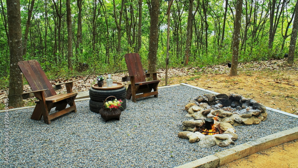 Two rock fire pit with wood table and chairs on gravel floor in the garden background. Stone fire pit with tire table decoration and wooden reclining chair on pebble ground. Two rock fire pit with wood table and chairs on gravel floor in the garden background. Stone fire pit with tire table decoration and wooden reclining chair on pebble ground.