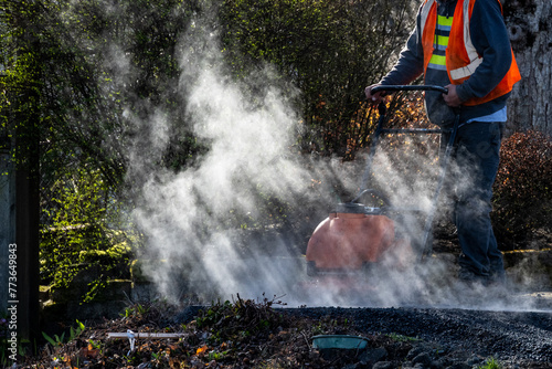 Construction worker in safety vest spraying using a rammer compactor to tamper down freshly laid asphalt at the end of a sidewalk with early morning sun shining through smoke and steam 