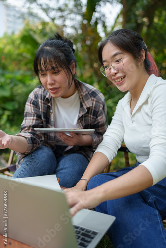 Two Asian female friends laugh and use tablets and laptops to study, learn, and research. Chat Online While relaxing on the grass in the green park holiday concept work lifestyle.