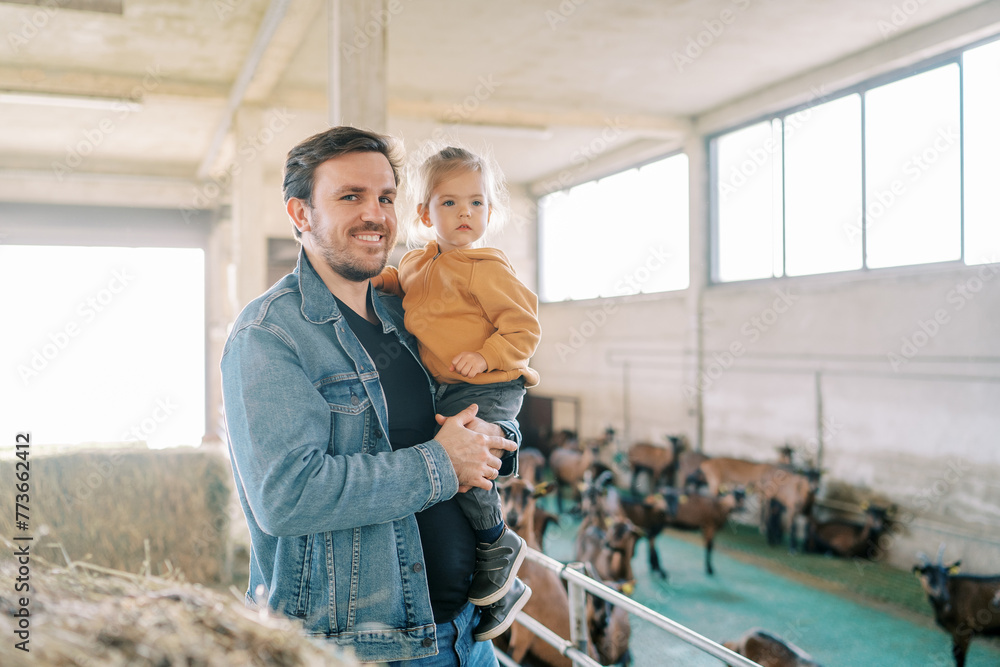 Smiling dad with a little girl in his arms stands at the pen with goats