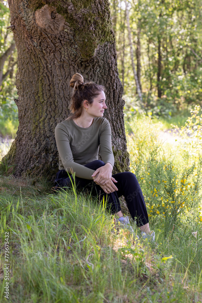 This image depicts a contemplative woman seated at the base of a large, textured tree in a verdant forest setting. Her gaze is directed off into the distance, suggesting introspection or a deep This image depicts a contemplative woman seated at the base of a large, textured tree in a verdant forest setting. Her gaze is directed off into the distance, suggesting introspection or a deep