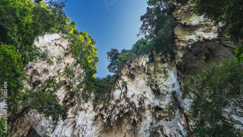 Green vegetation grows on the steep slopes of the cliff. The clear blue sky is visible through the hole from the bottom of the cave. Frame. Malaysia, Kuala Lumpur. Batu Caves