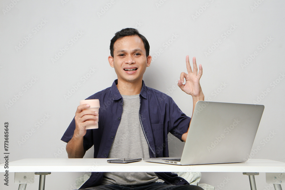 joyfull happy asian man at workplace holding a cup while sitting in front of laptop computer. on isolated background joyfull happy asian man at workplace holding a cup while sitting in front of laptop computer. on isolated background